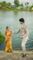 A couple by a lake during a wedding photo shoot; the man in a beige suit offers his hand to the woman in a gold and red sari by the water.