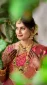 Portrait of an Indian bride in a red and gold sari wearing heavy gold jewelry and a maang tikka, with hennaed hands posed at chest level amid greenery