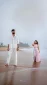 Man in white linen outfit standing barefoot on a beach with a woman in a pastel dress walking near the water behind him.
