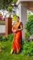 Woman in a bright orange and gold saree, wearing heavy jewelry, standing in a garden beside a column.
