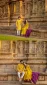 Couple in yellow traditional attire sits by an ancient carved stone wall with figures.