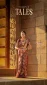 Woman in a maroon and gold saree with heavy jewelry standing beside an ornate carved stone wall at a temple entrance, posing for a photo.