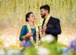 Bride and groom in traditional blue outfits wearing flower garlands, smiling at each other at their wedding ceremony while decorative lights glow behind them.