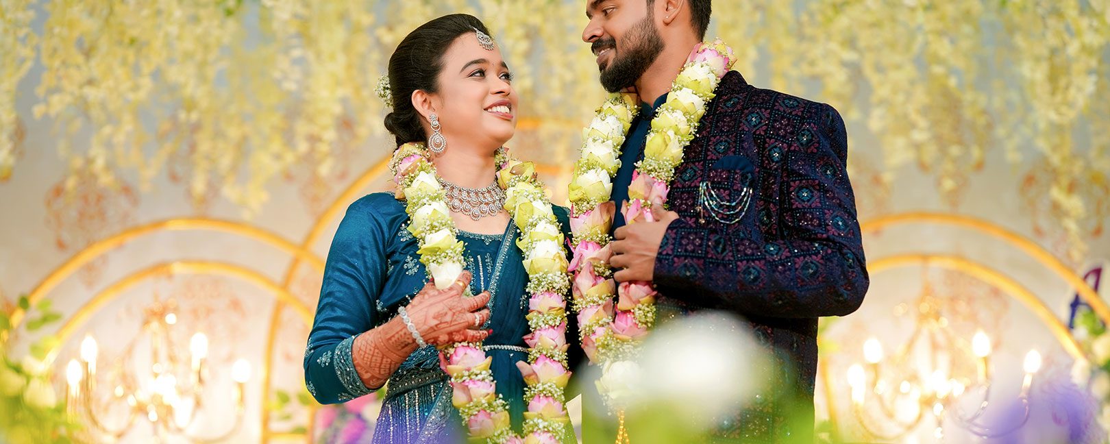 Bride and groom in traditional blue outfits wearing flower garlands, smiling at each other at their wedding ceremony while decorative lights glow behind them.