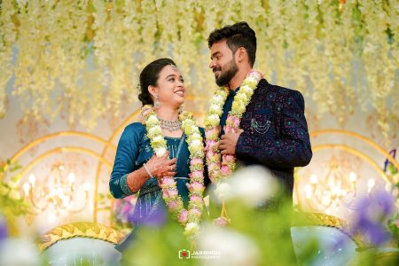 Bride and groom in traditional blue outfits wearing flower garlands, smiling at each other at their wedding ceremony while decorative lights glow behind them.