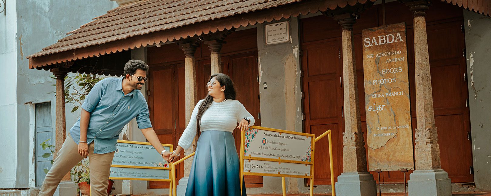 Young couple holding hands and smiling in front of a storefront with a SABDA sign and orange barriers nearby.