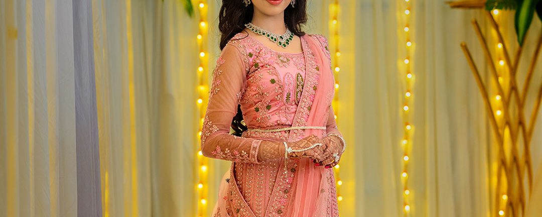 Woman in a pink embellished lehenga standing amid warm string lights and floral decor at a festive event, posing with hands clasped.