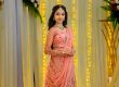 Woman in a pink embellished lehenga standing amid warm string lights and floral decor at a festive event, posing with hands clasped.