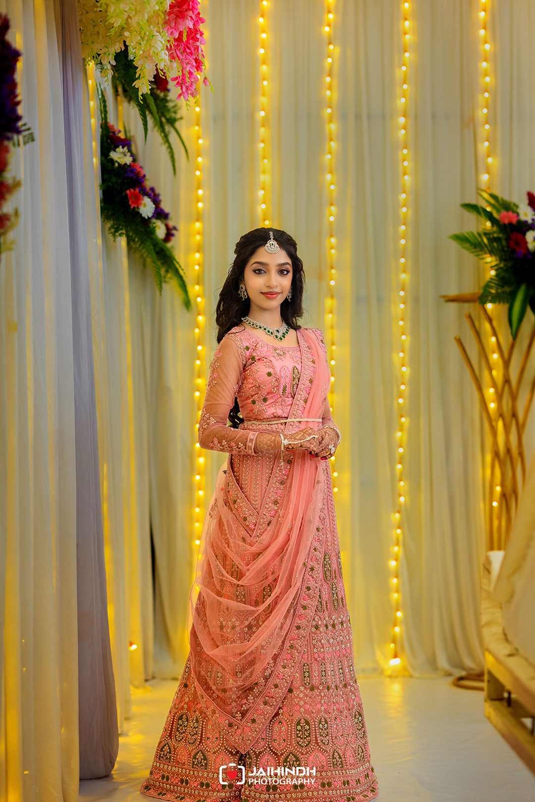 Woman in a pink embellished lehenga standing amid warm string lights and floral decor at a festive event, posing with hands clasped.