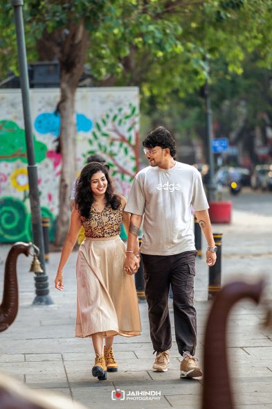 Young couple walking hand in hand on a city sidewalk, smiling, with a colorful mural in the background read more of the wall art behind them.