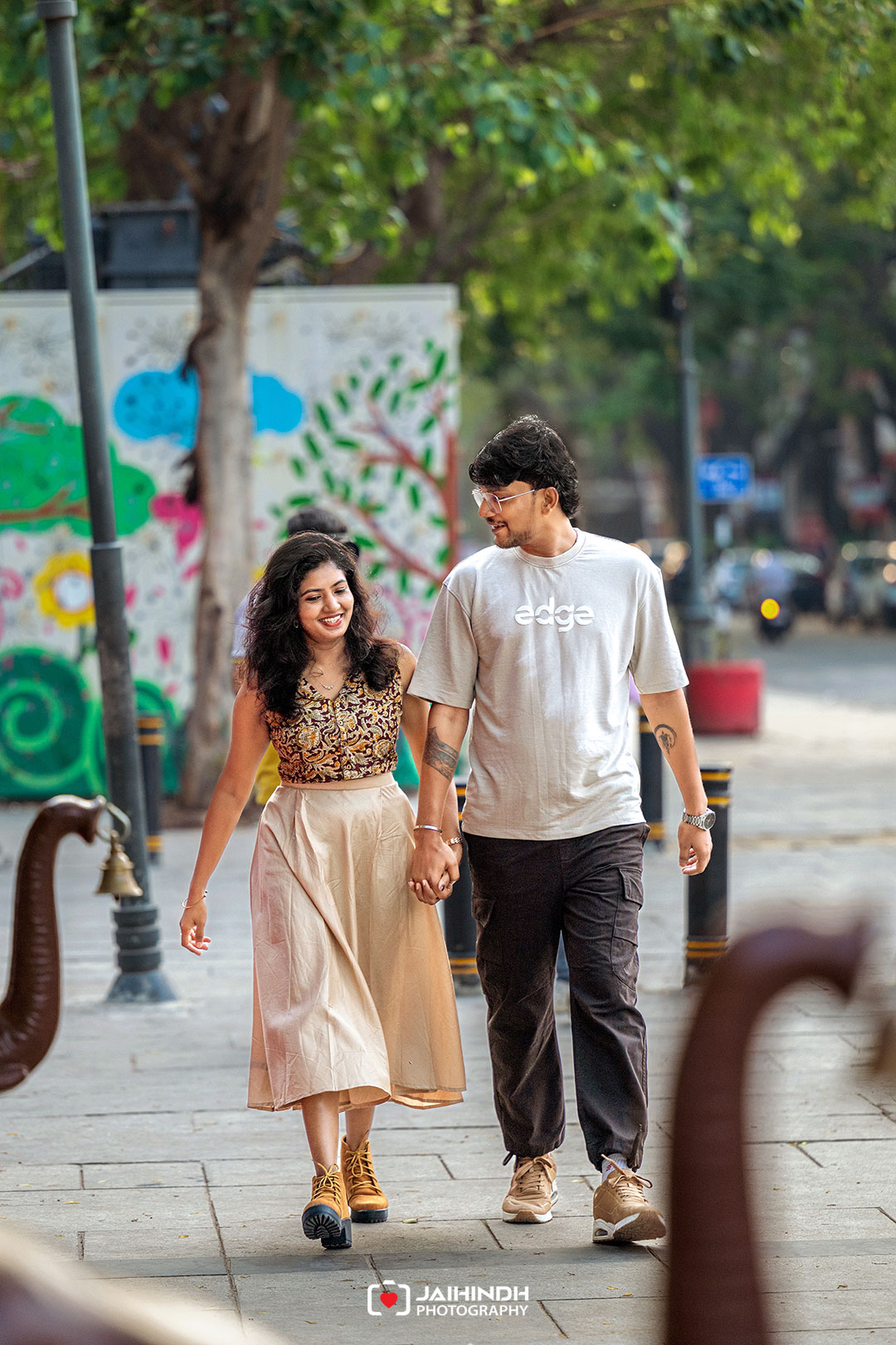 Young couple walking hand in hand on a city sidewalk, smiling, with a colorful mural in the background read more of the wall art behind them.
