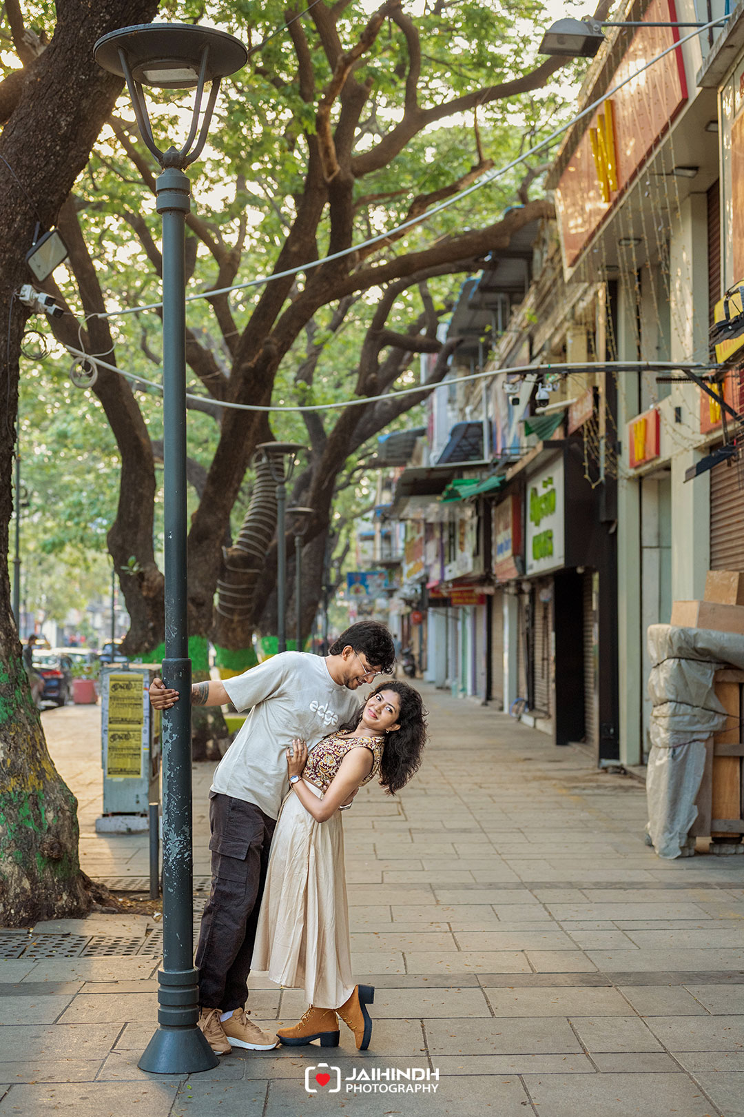 Couple leaning on a tall streetlamp and sharing a moment on a tree-lined city sidewalk with shops in the background.