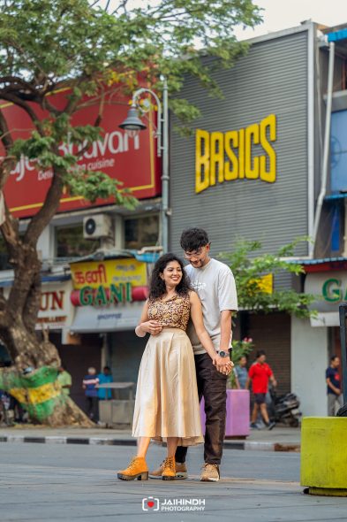 A smiling couple stands close together on a city sidewalk, holding hands with colorful storefronts and signs in the background.