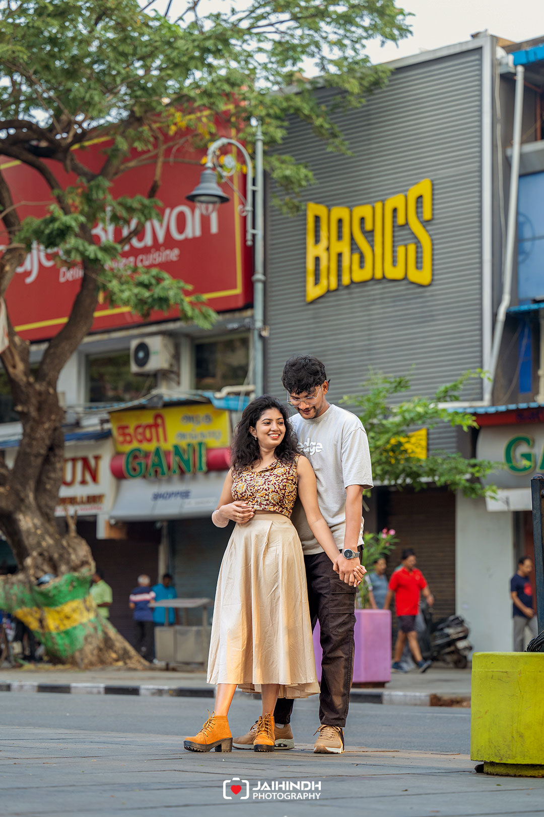 A smiling couple stands close together on a city sidewalk, holding hands with colorful storefronts and signs in the background.