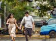 Young couple holding hands and running along a city street with pigeons flying overhead and green trees above.