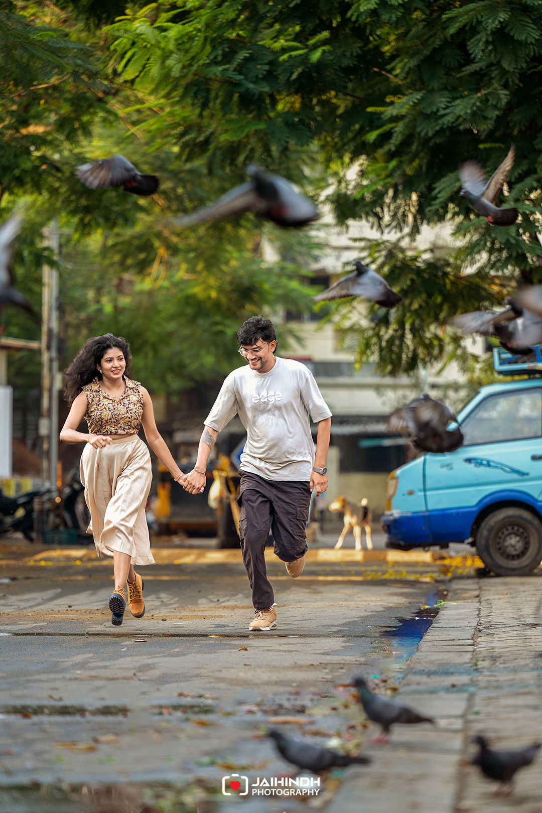 A couple holding hands and running down a street as pigeons fly around them under green trees.