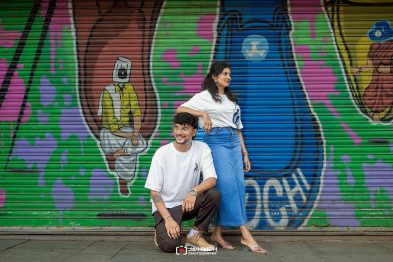 Two friends pose in front of a vibrant graffiti mural; a man kneels while a woman stands beside him, smiling.