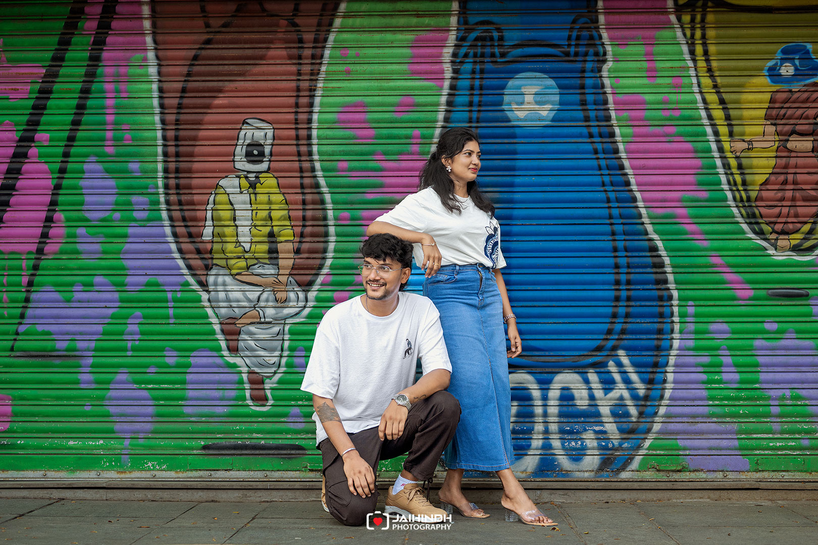 Two friends pose in front of a vibrant graffiti mural; a man kneels while a woman stands beside him, smiling.