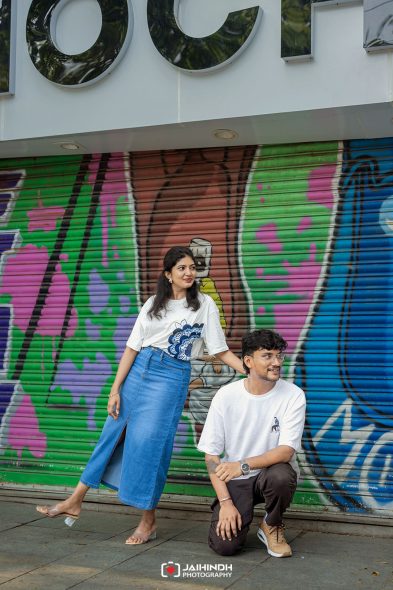 Woman in a white graphic tee and blue skirt poses beside a kneeling man in front of a colorful graffiti wall.