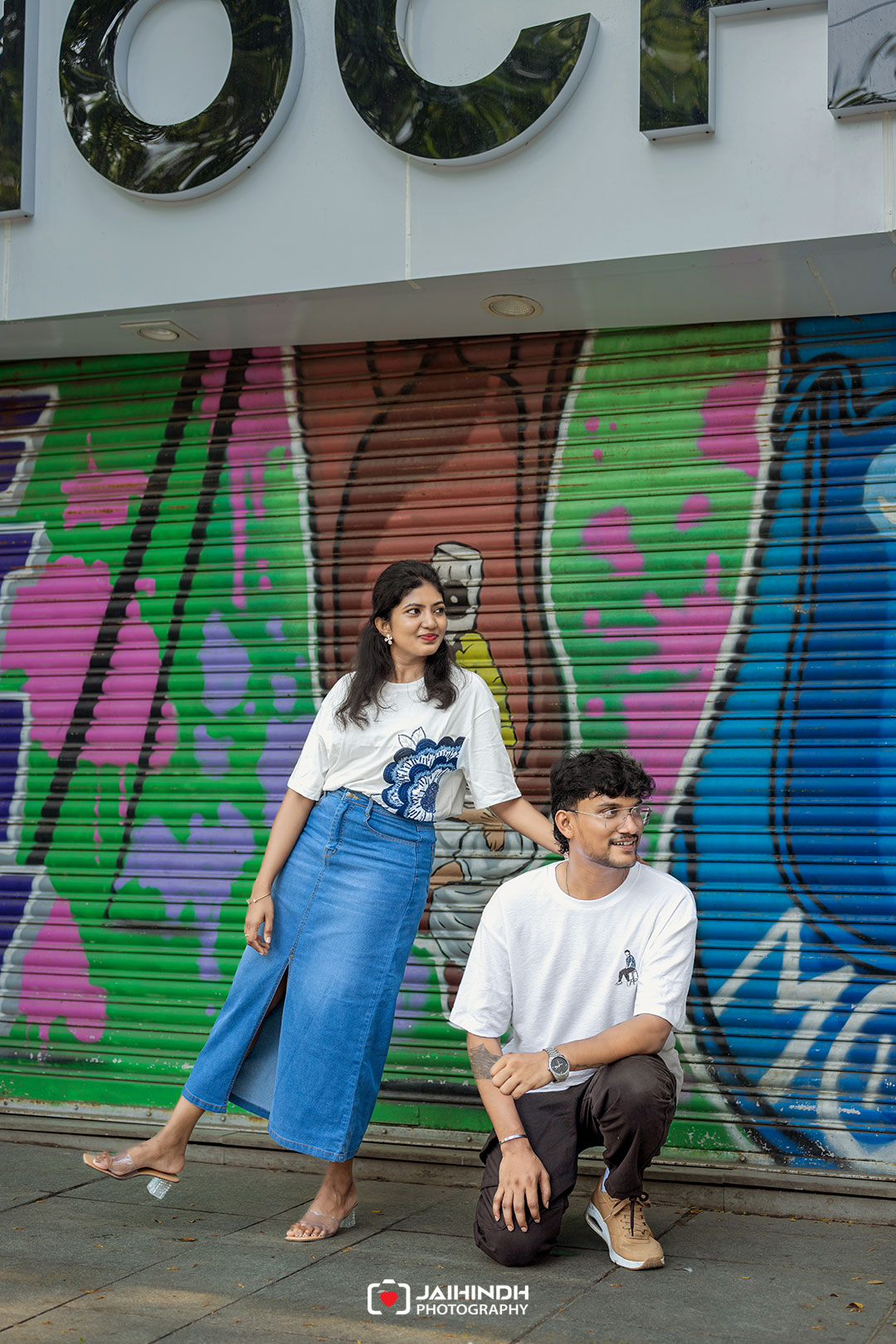 Woman in a white graphic tee and blue skirt poses beside a kneeling man in front of a colorful graffiti wall.