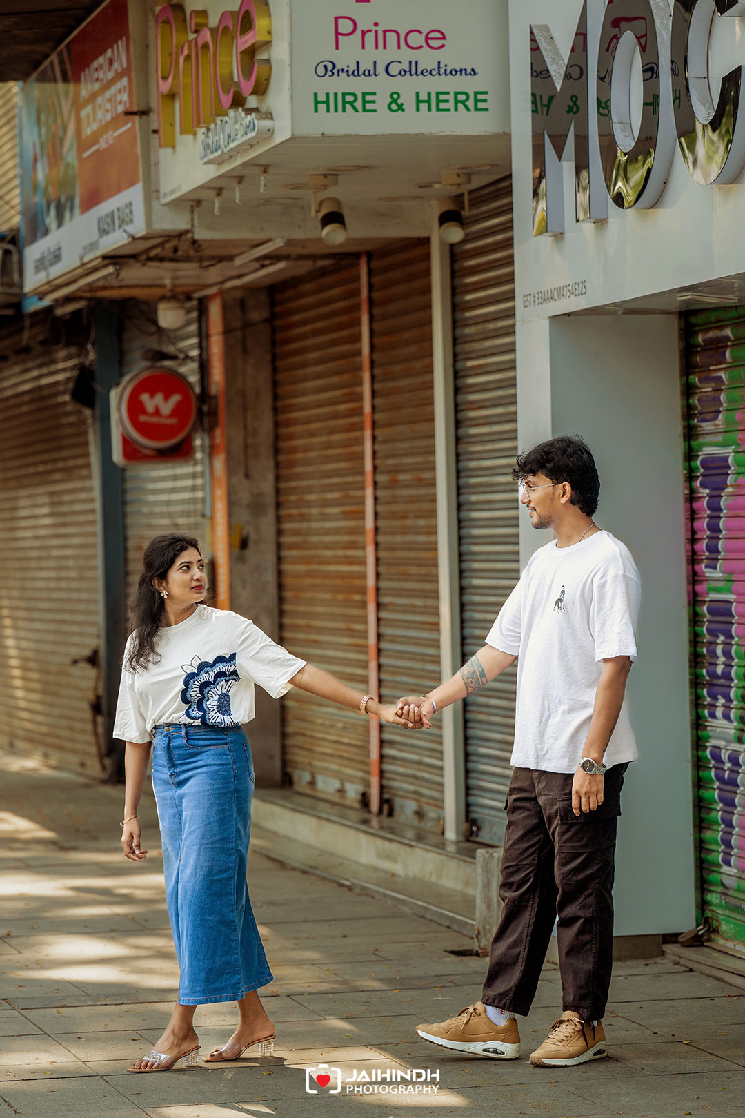 Young couple holding hands while walking down a sunny sidewalk in an urban shopping street with closed storefronts behind them.