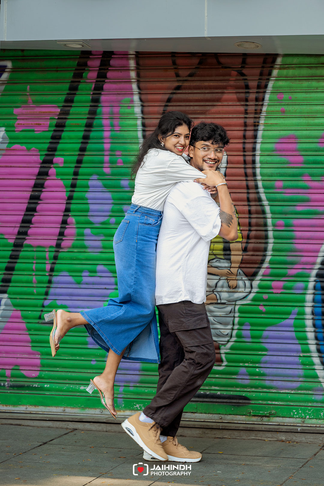 A smiling couple hugs in mid-air in front of a colorful graffiti wall.
