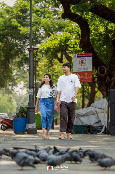 Couple walking hand-in-hand along a city sidewalk with pigeons in the foreground and green trees in the background, smiling at each other.