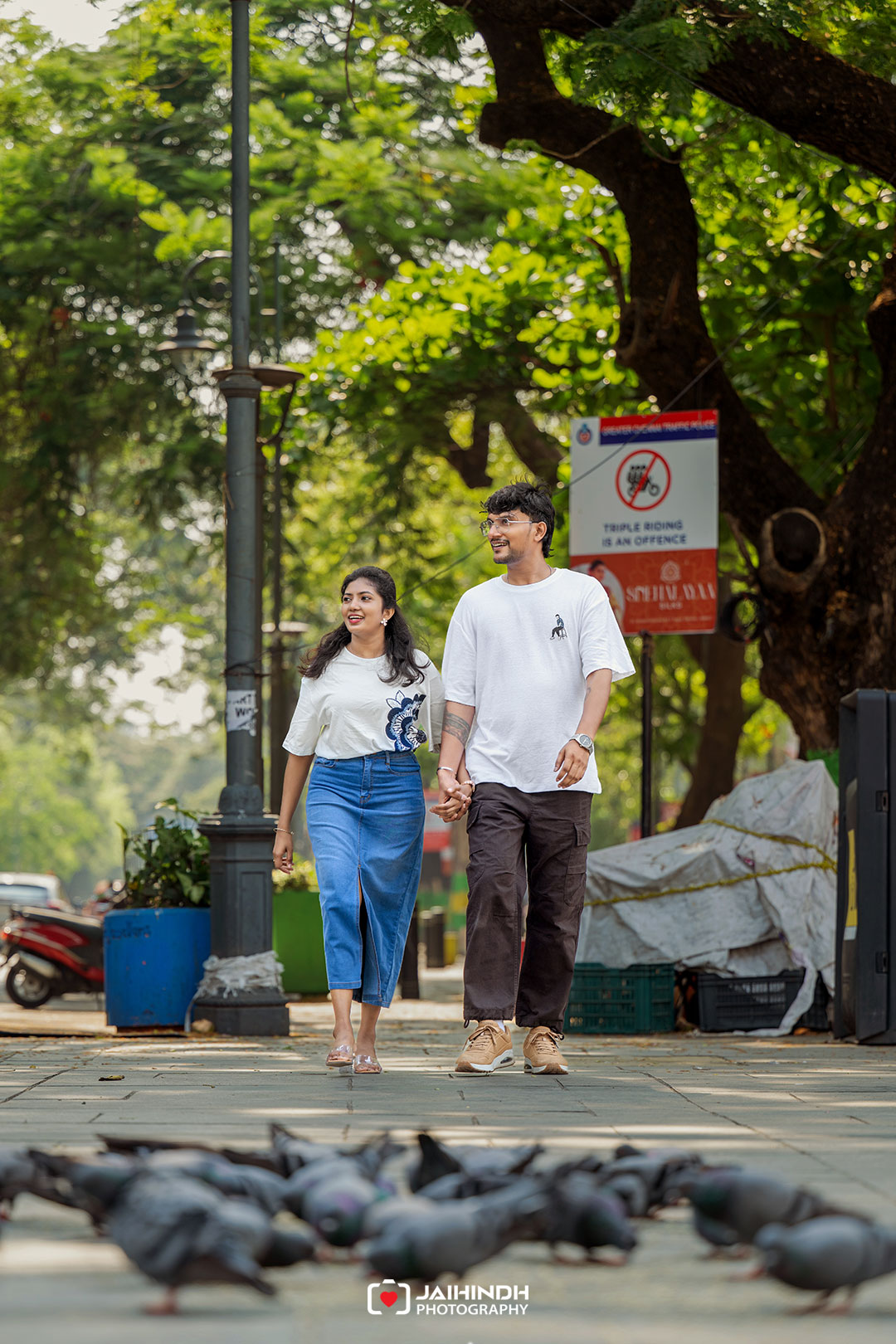 Couple walking hand-in-hand along a city sidewalk with pigeons in the foreground and green trees in the background, smiling at each other.