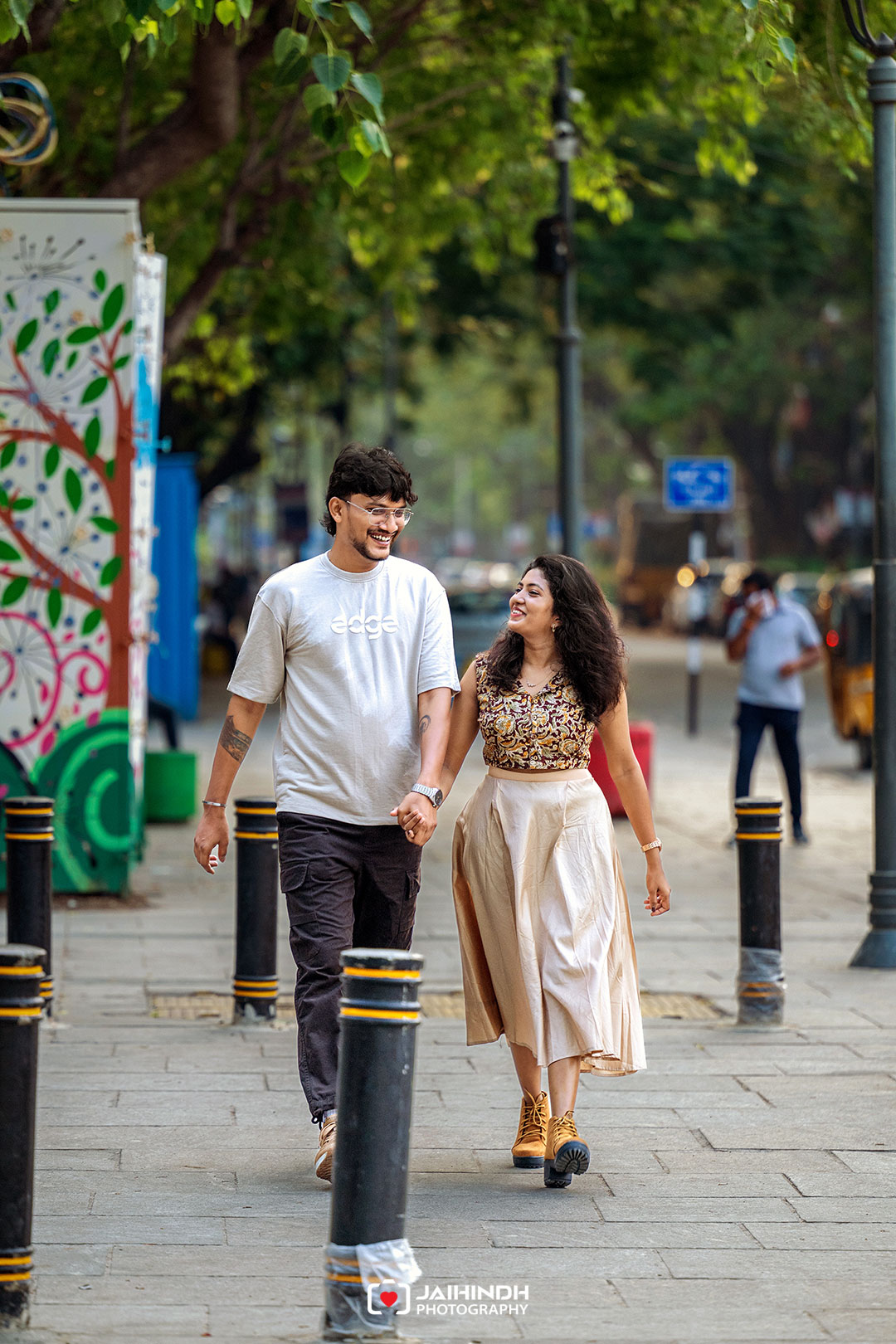 Couple holding hands and smiling while walking down a street with a colorful mural on the left and bollards along the sidewalk.