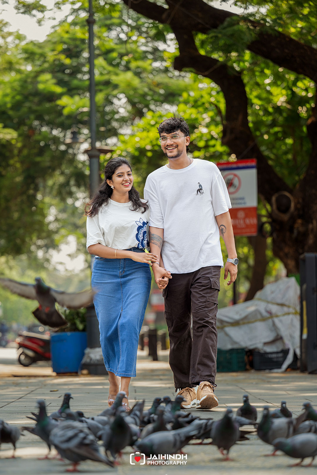 A young couple walks hand in hand along a park path, smiling, with pigeons on the ground in front and green trees in the background.