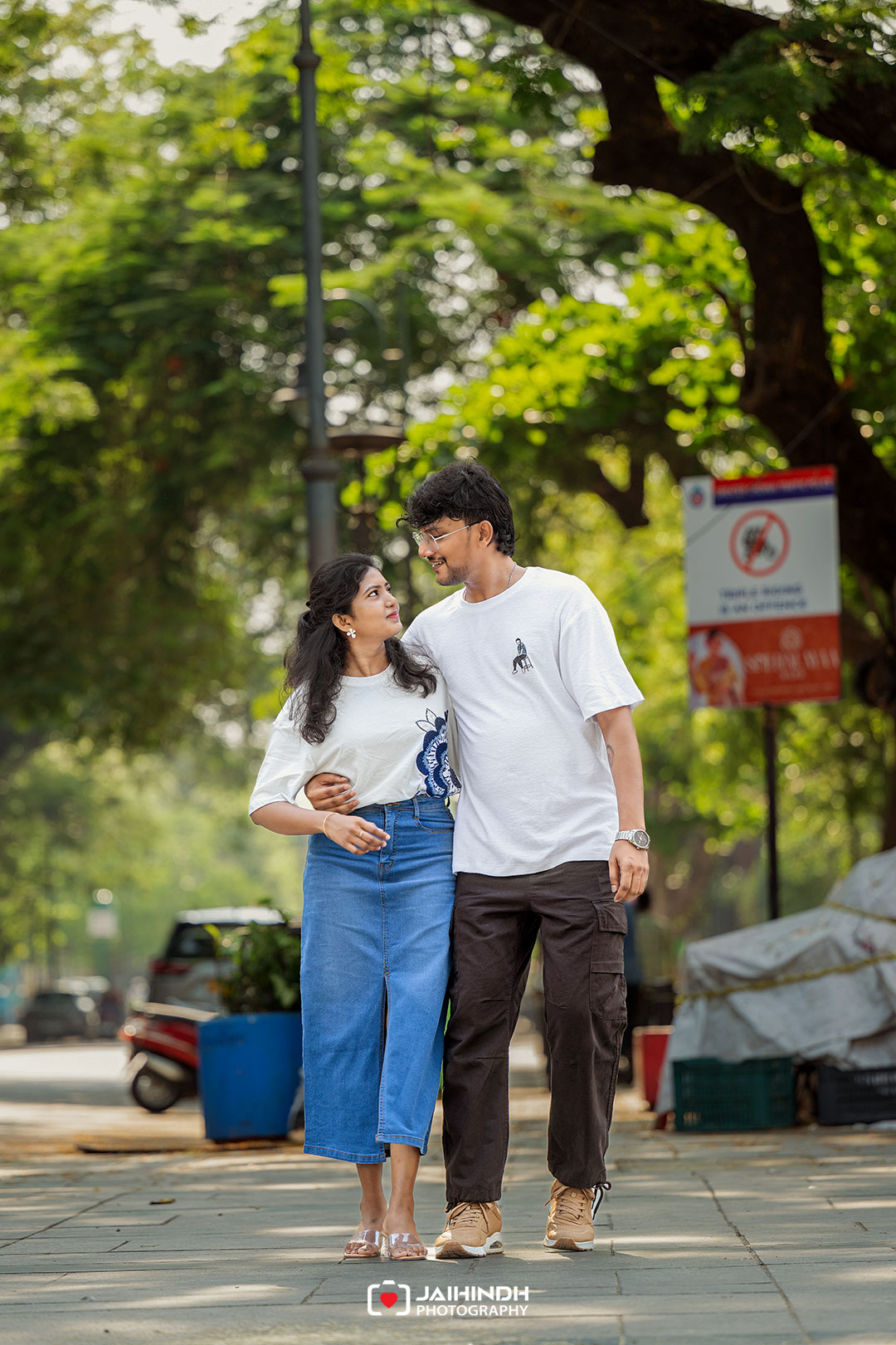 A couple walks arm-in-arm on a city sidewalk, smiling at each other with green trees in the background.