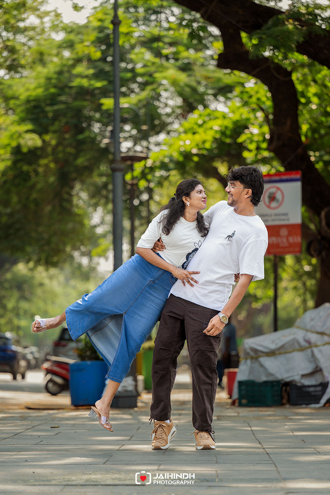 A young couple smiles at each other as the man lifts the woman playfully on a park walkway surrounded by trees.