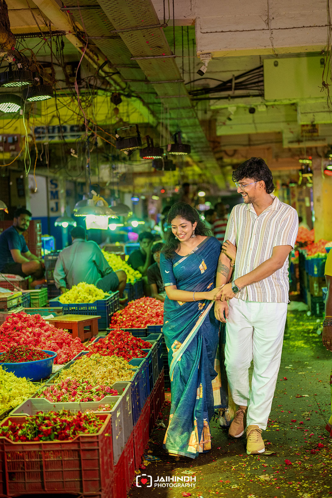 Smiling couple strolling through a busy night market with colorful flower and vegetable crates; woman in a blue sari, man in a striped shirt.