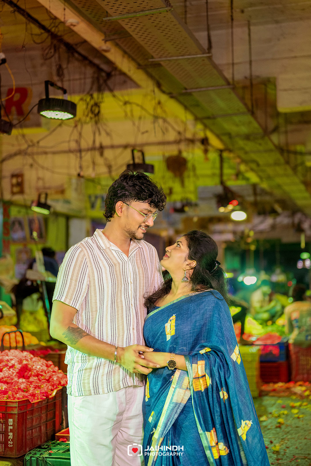 Couple stands close in a busy indoor market, holding hands and gazing at each other; fruit stalls and green lighting surround them.
