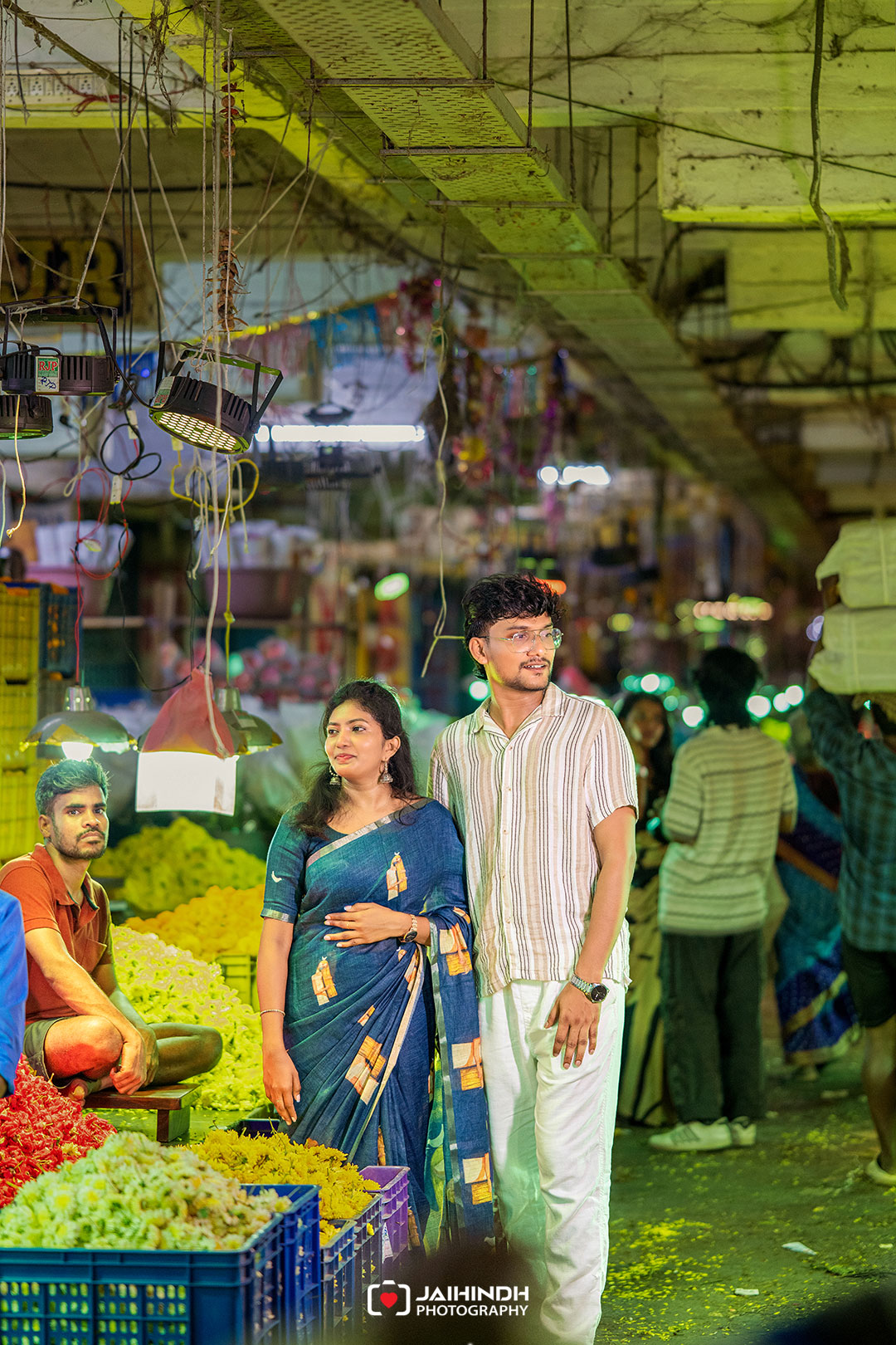 Couple in a busy indoor market standing beside crates of colorful vegetables, with vendors and other shoppers in the background.