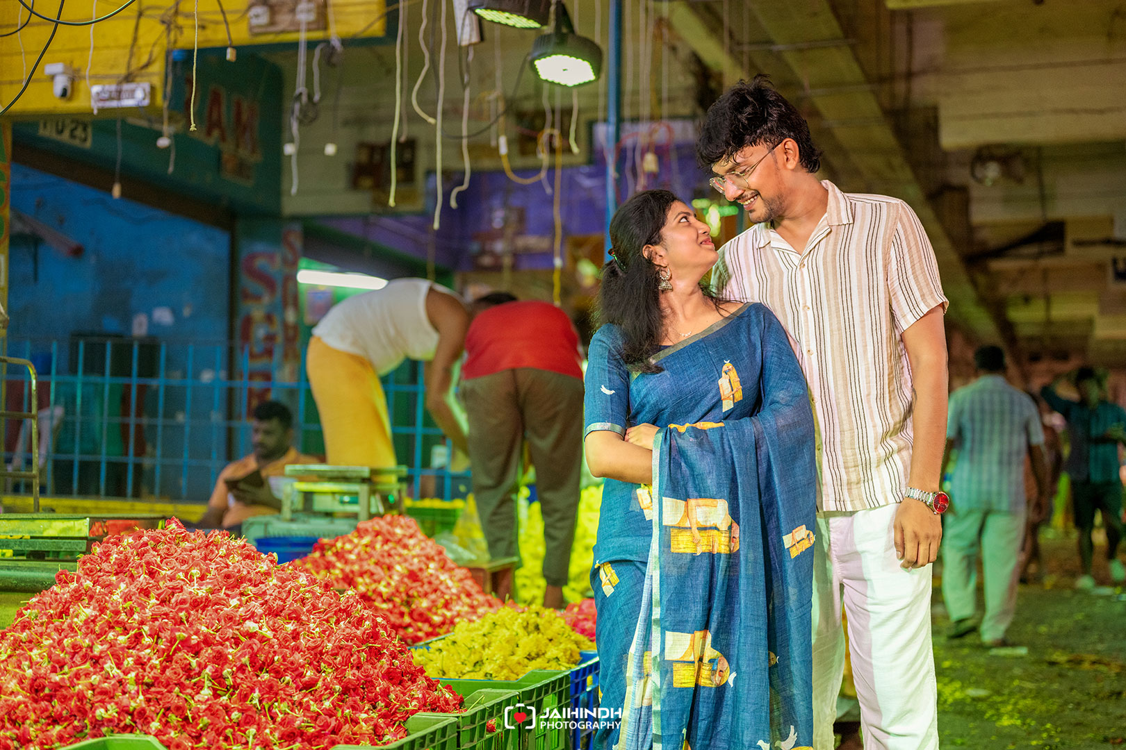 Couple smiling at each other in a crowded flower market at night, with red and yellow flowers in the foreground.