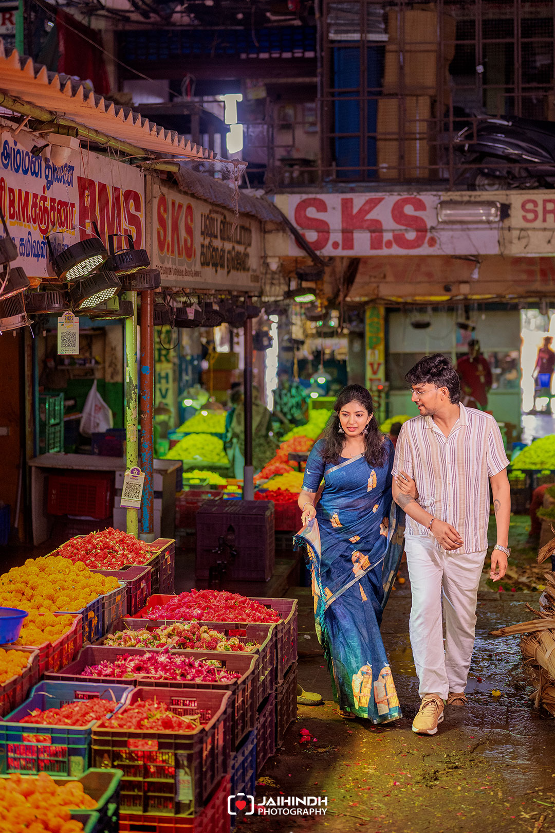 A couple walks arm in arm through a colorful market street with crates of yellow, pink, and red produce nearby.