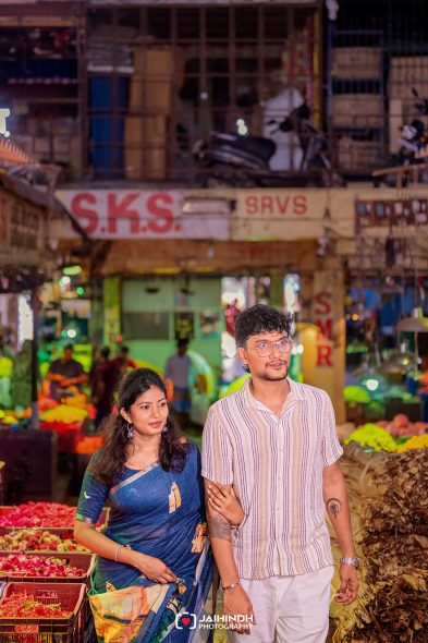 A young couple walks arm in arm through a busy night market with colorful fruit and vegetables around them.