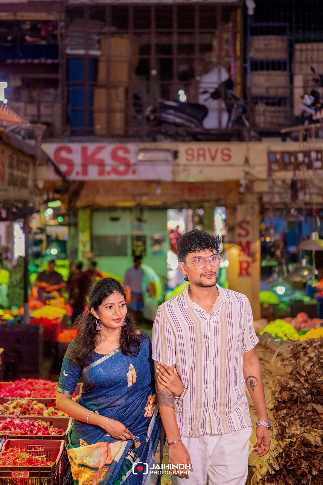 A young couple walks arm in arm through a busy night market with colorful fruit and vegetables around them.