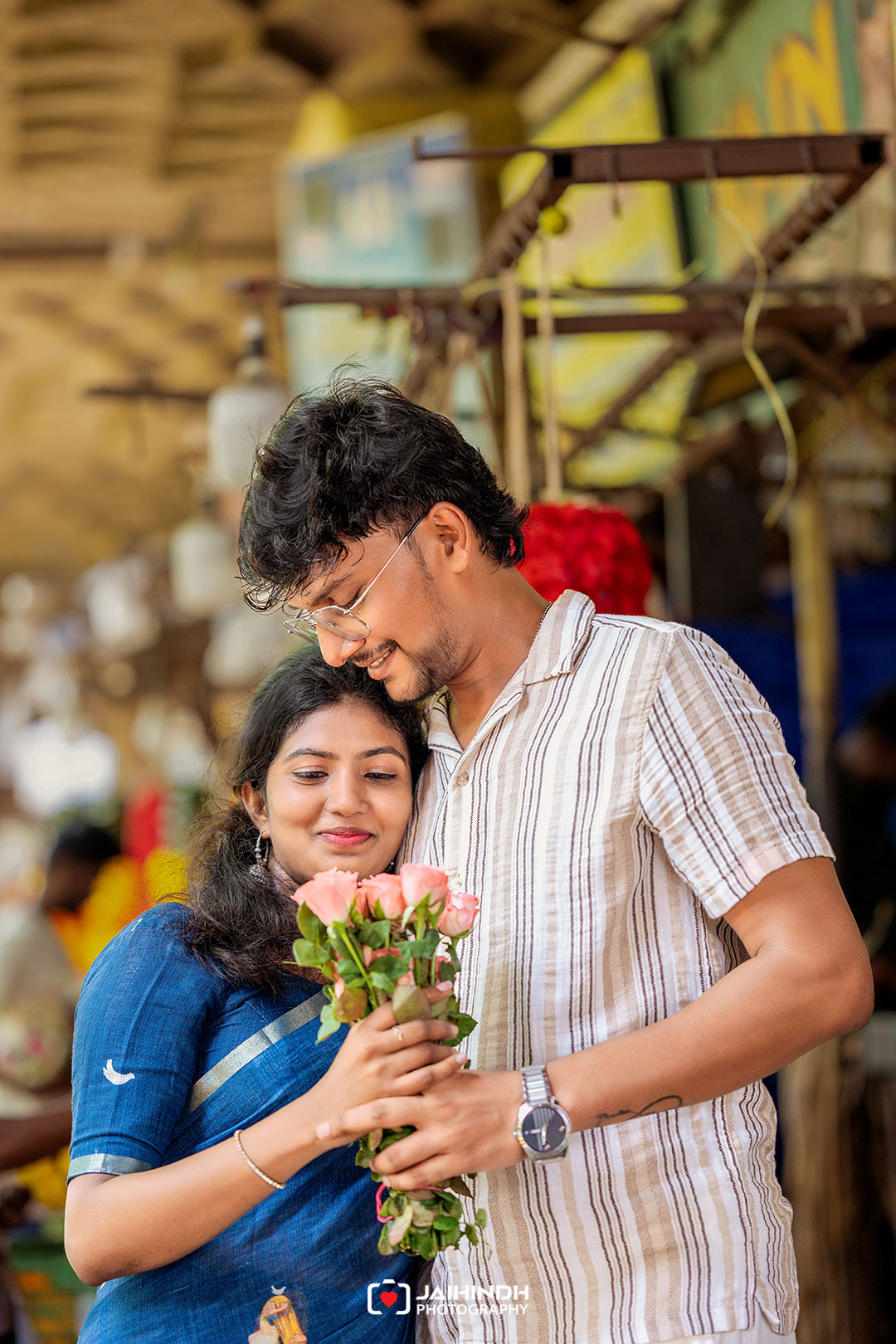 Couple in a market sharing a tender moment; man hugs a woman while they hold a bouquet of pink roses.