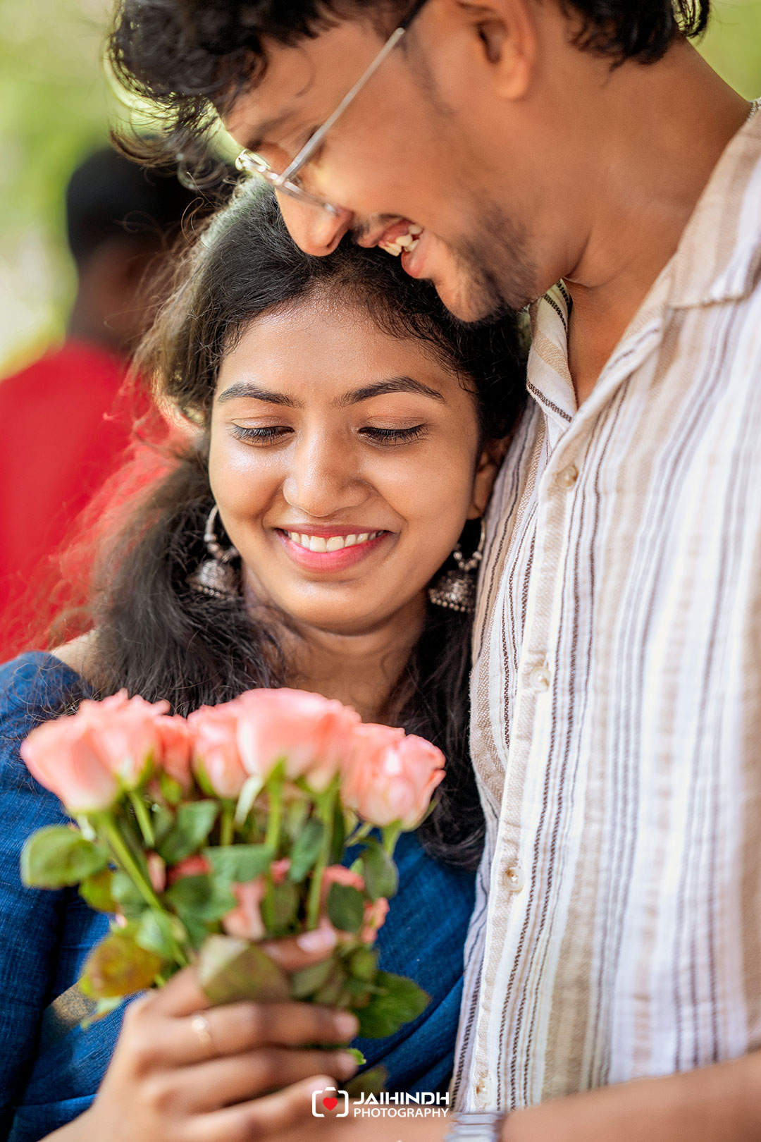 Smiling couple embracing outdoors, woman holding a bouquet of pink roses.