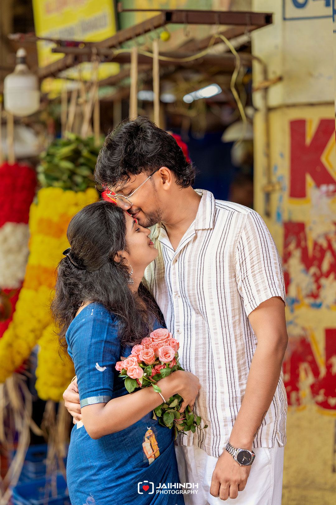 Couple sharing a gentle kiss in a colorful market, the woman holding pink roses while the man wears a striped shirt; vibrant stalls in the background.