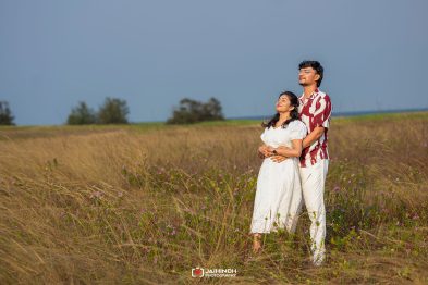 Couple standing in a tall grassy field, embracing and basking in sunlight with a blue sky backdrop.
