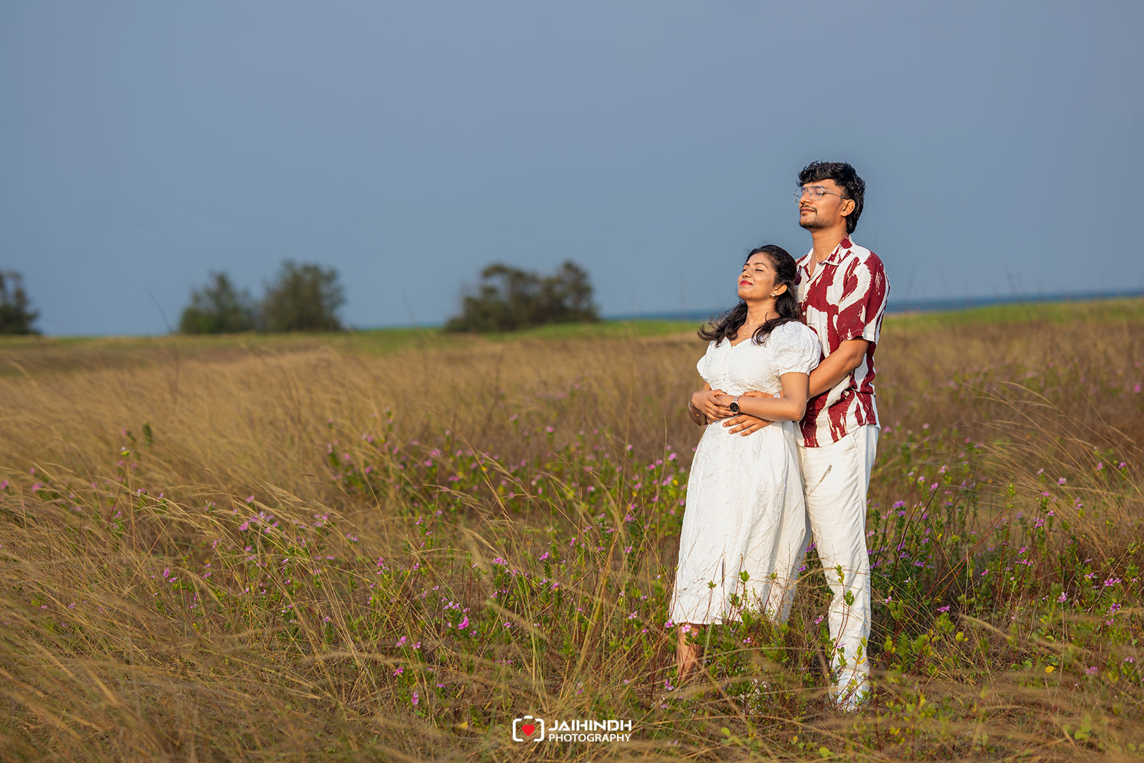 Couple standing in a tall grassy field, embracing and basking in sunlight with a blue sky backdrop.