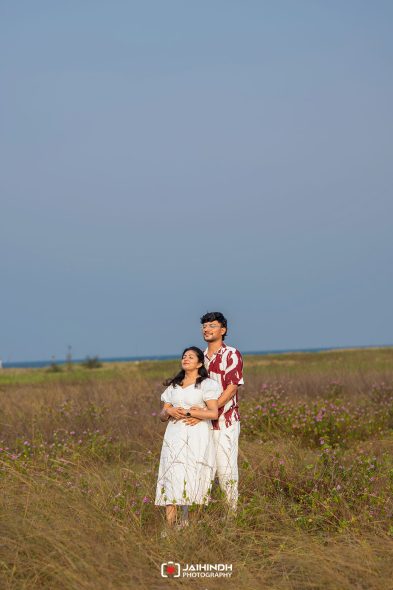 Couple standing in a sunlit field of tall grasses and pink flowers, with the ocean and a clear blue sky in the background.