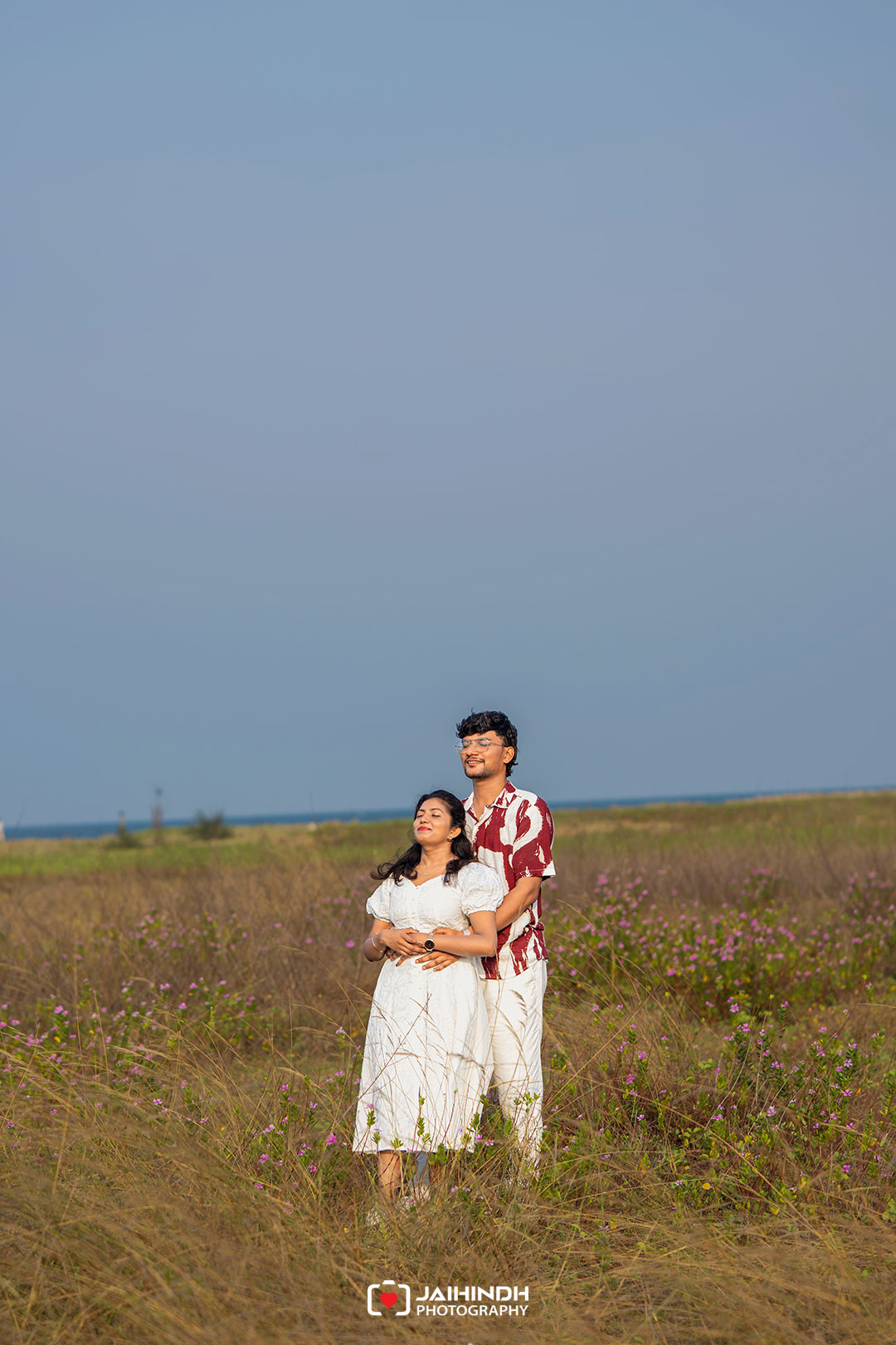 Couple standing in a sunlit field of tall grasses and pink flowers, with the ocean and a clear blue sky in the background.