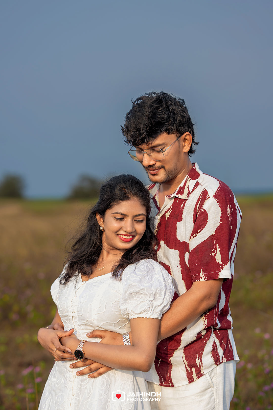 A couple stands in a sunlit field; the man embraces the woman from behind as they smile together.