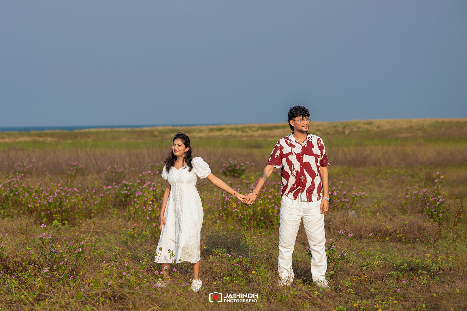 Couple holding hands in a meadow of pink wildflowers with the sea on the horizon under a clear blue sky.