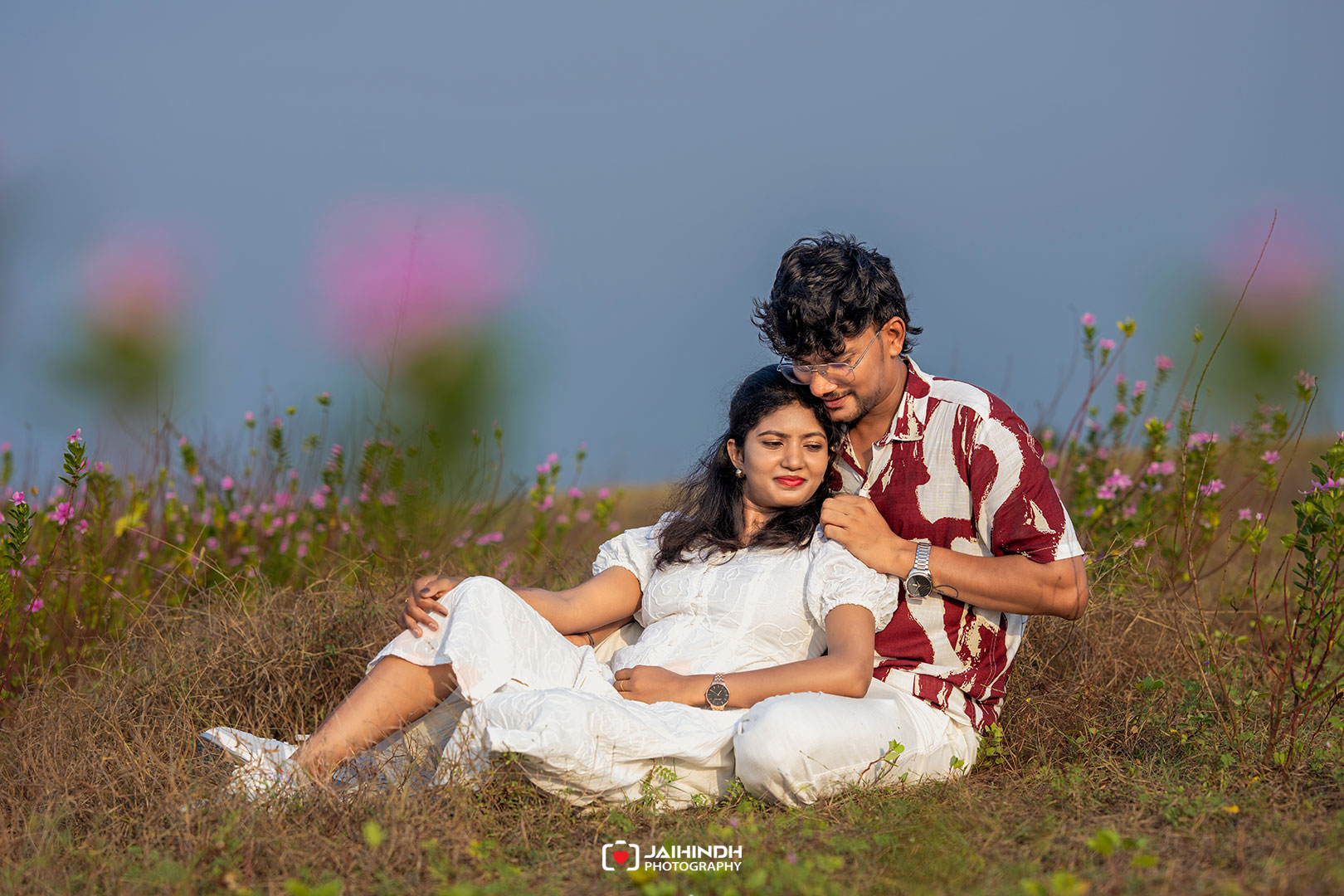 A couple sits in a field of wildflowers, the man embracing the woman as they smile together in the sun.