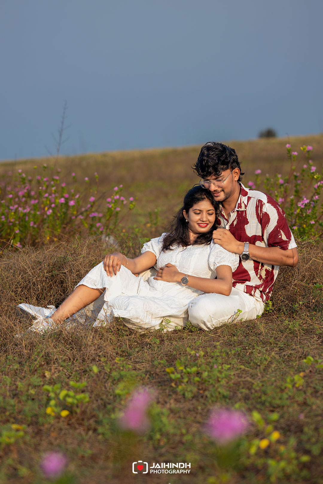 Couple sitting on the grass in a sunny meadow of pink flowers, the woman in white leaning on the man in a red-and-white shirt, both smiling affectionately.